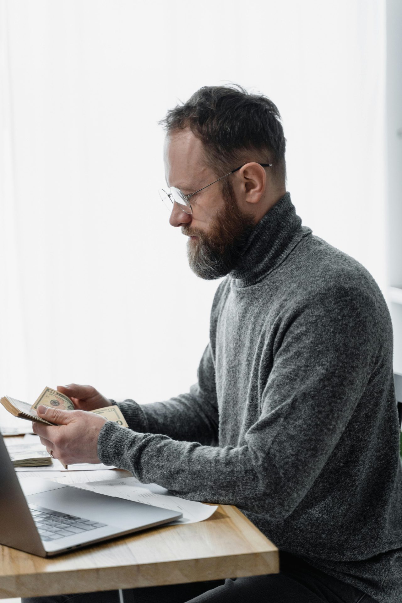 A man counts cash while working on a laptop at his desk in a modern office setting.