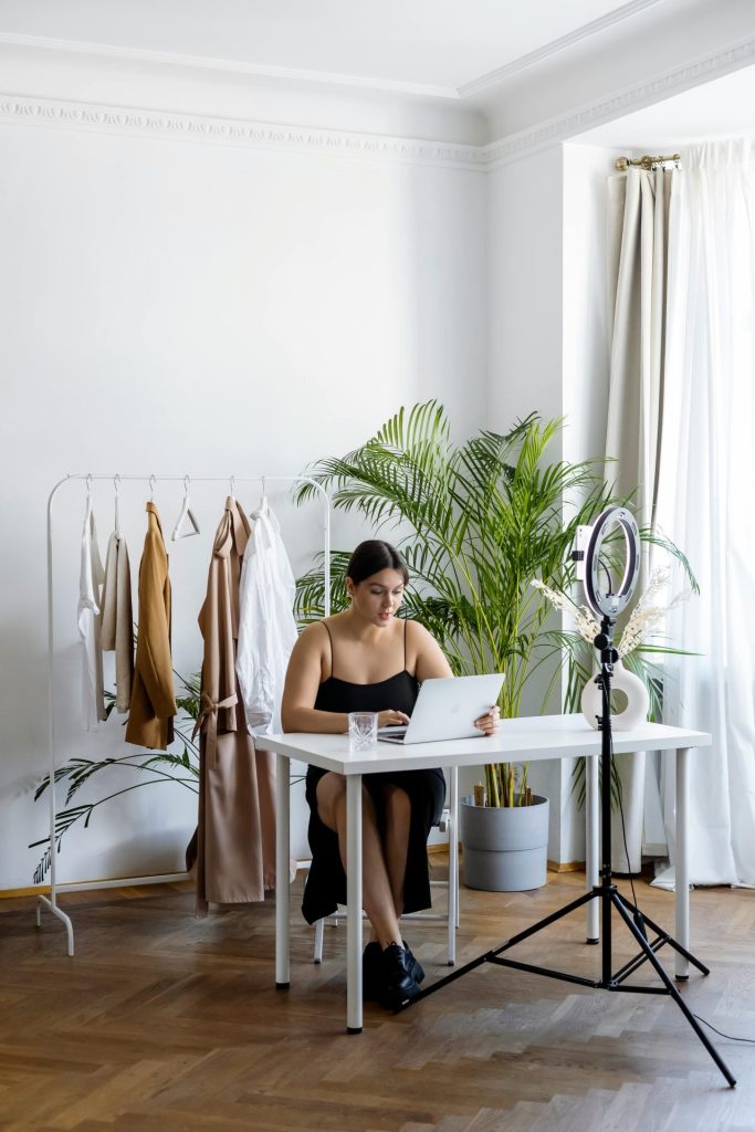 A woman engaging in online selling with clothes rack, laptop, and ring light setup indoors.