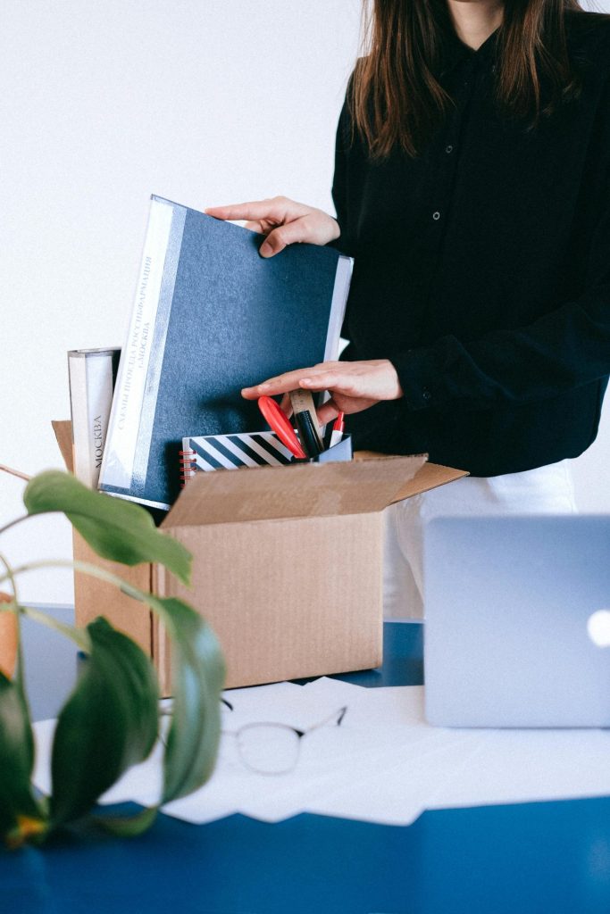 Adult woman organizing office supplies into a cardboard box, possibly symbolizing job change or relocation.