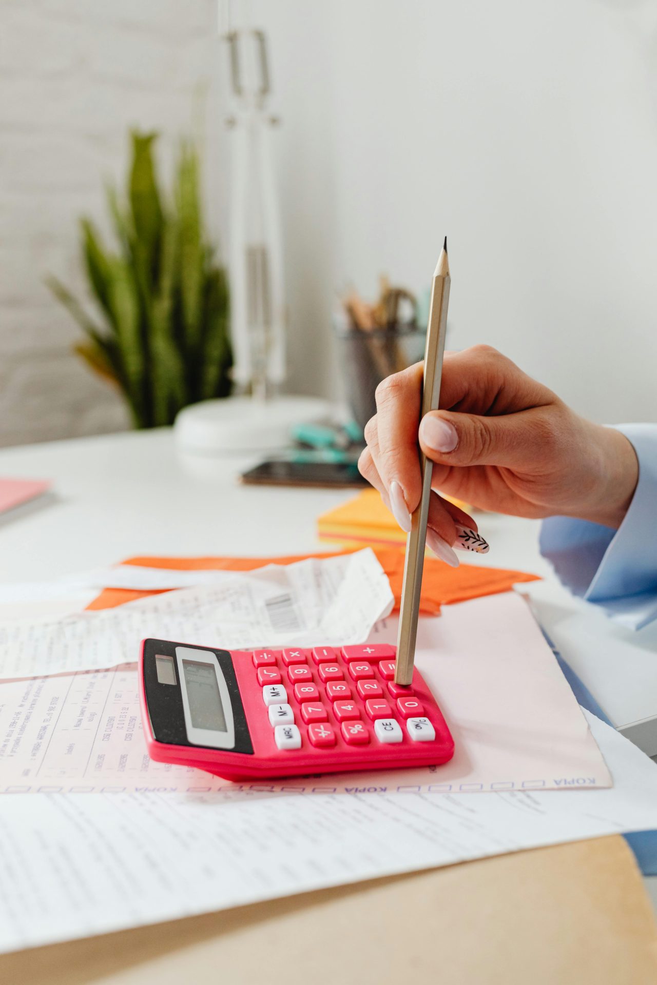 Close-up of a hand using a pencil with a red calculator, surrounded by papers indoors.